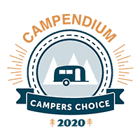 A circular logo with "Campendium Campers Choice 2020" text. The logo features an illustration of a camper trailer against a backdrop of mountain peaks, bordered by a stitched pattern and stylized orange rays, celebrating the Best RV Park with the Campers Choice Award.