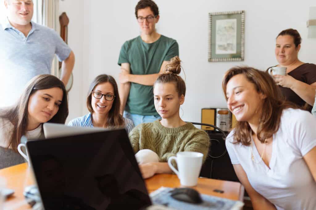 A group of seven people are gathered around a table, smiling and looking at a laptop screen. The table has coffee mugs and papers on it. The room appears bright and casual with a "Now Hiring" sign on the wall. One person stands behind the others with a cup, observing.