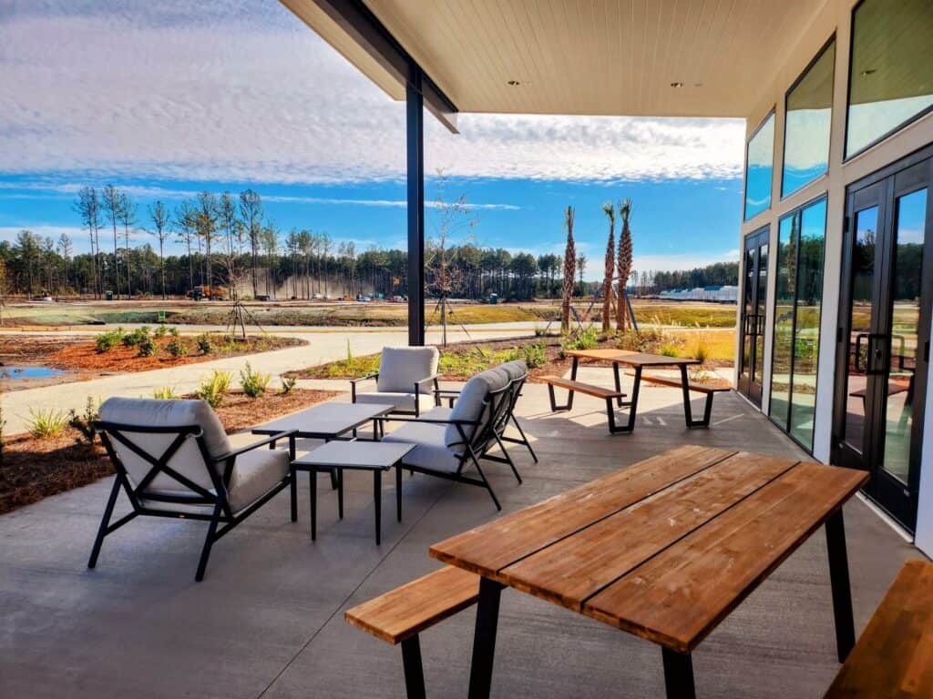 Outdoor patio with modern furniture under a sheltered roof, overlooking a scenic landscape with trees and open fields. Wooden picnic tables and cushioned seating arrangements are placed on the concrete floor, creating a serene and inviting atmosphere perfect for weekly gatherings or monthly events.