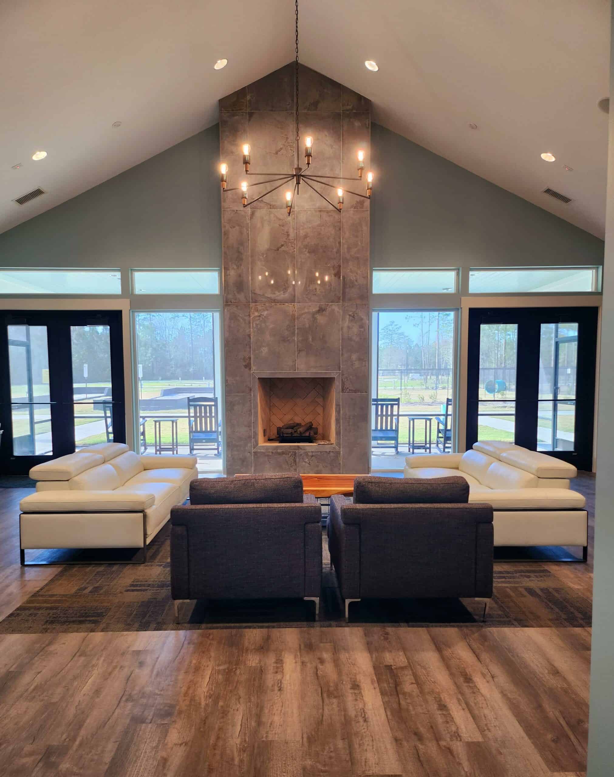 A modern living room with a vaulted ceiling, featuring a large stone fireplace at the center. The room has two white leather couches and two dark armchairs facing the fireplace, along with a chandelier above. Large windows and glass doors allow natural light to flood the space.