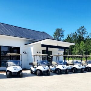 A row of eight white golf carts is parked neatly in front of a modern white building with large windows. The scene is set under a clear blue sky, with trees visible in the background. The building has a dark metal roof and a small overhang above the entrance.