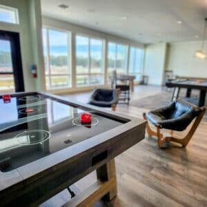 A modern recreational room with large windows offering a scenic view. The room features various game tables, including an air hockey table in the foreground. There are comfortable chairs and tables scattered throughout the space, creating a relaxed atmosphere.