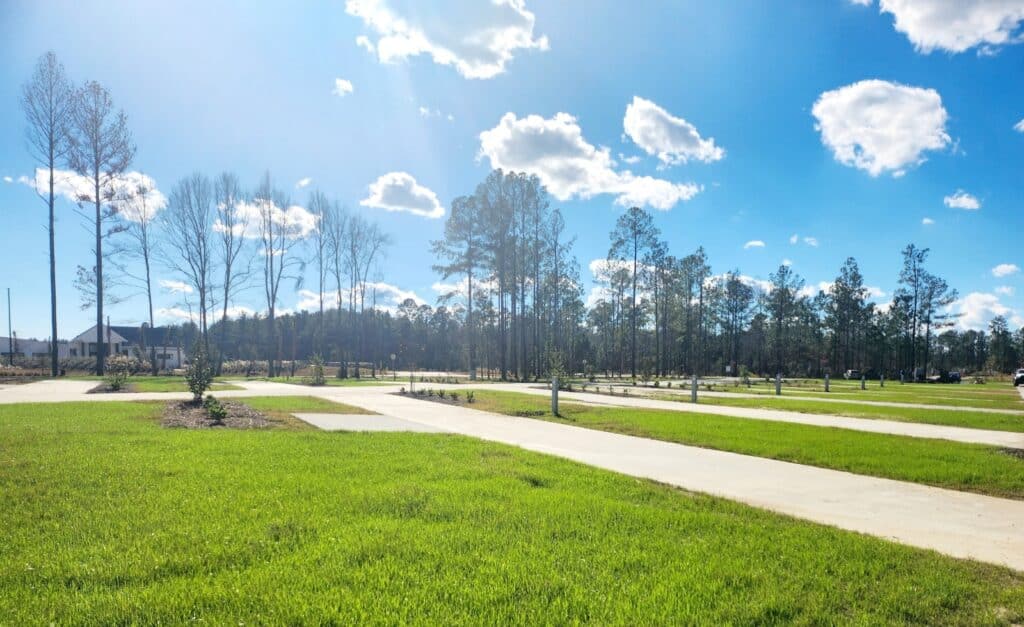 A serene park with green grass fields and neatly paved pathways under a bright blue sky with scattered clouds. Tall, leafless trees line the park, overlooking RV camping sites. A dense forest can be seen in the background, with distant structures visible beyond the trees.