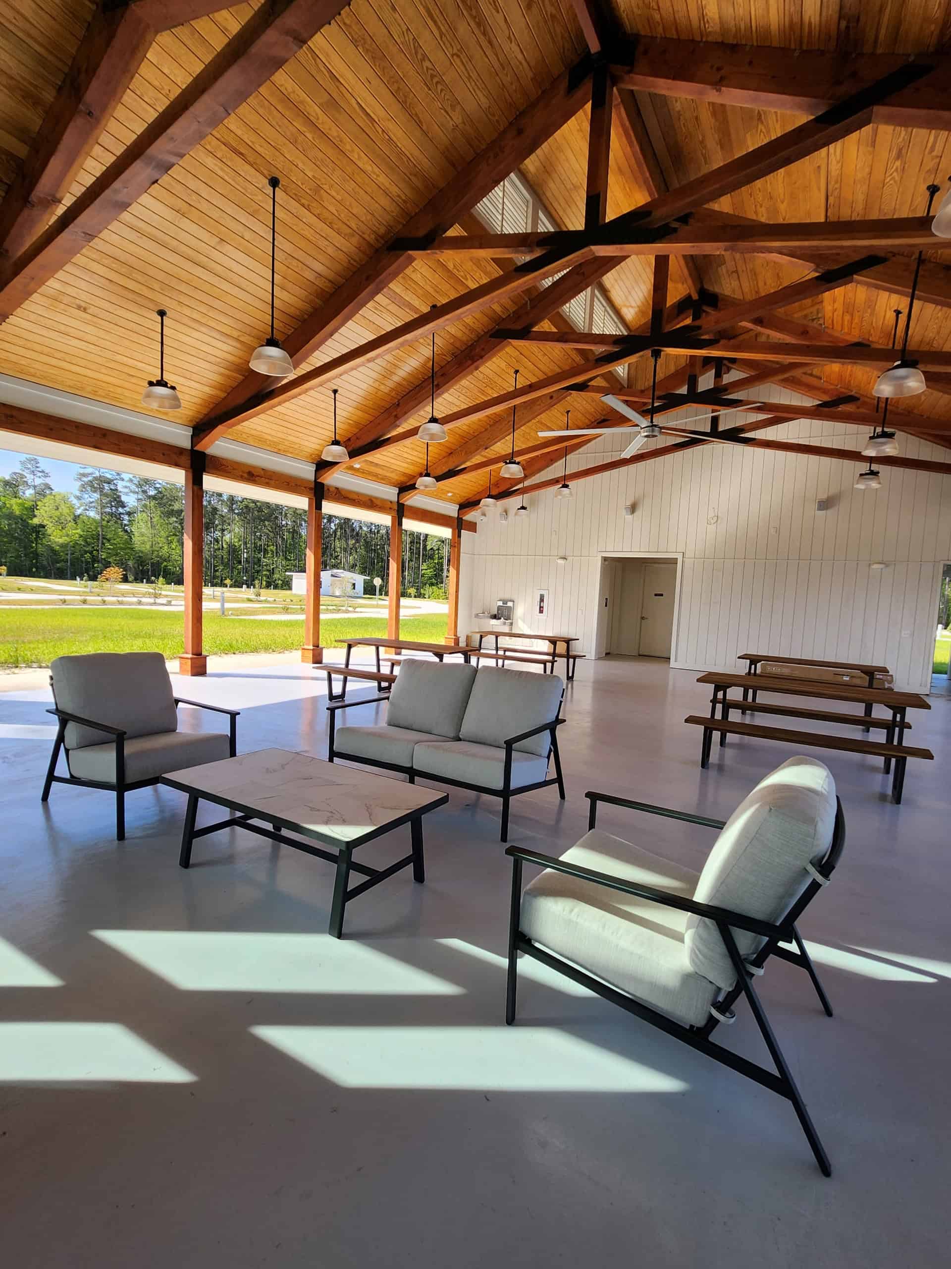 A spacious, open-air pavilion with a wooden ceiling, pendant lights, and ceiling fans. Inside, there is a seating area with cushioned chairs and coffee tables on one side, and picnic tables on the other. Large windows provide a view of a grassy field with trees.