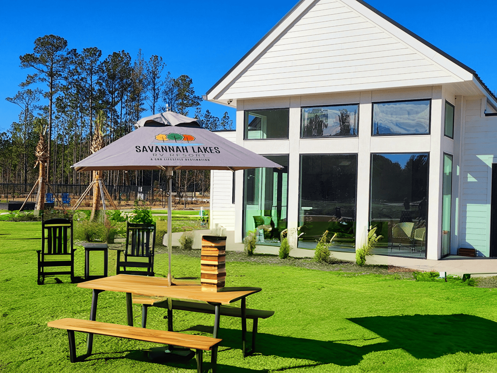 Modern house with large windows and manicured lawn. A picnic table with benches is topped by a large umbrella labeled "Savannah Lakes." Tall green trees line the background under a clear blue sky. Three Adirondack chairs are arranged around the lawn.