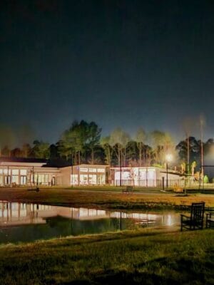 A serene nighttime scene at the Savannah Lakes RV Resort features a well-lit building with large windows, surrounded by trees. In the foreground, a tranquil pond reflects the building's lights, and a bench sits on the grass near the water's edge. The sky is dark, and the surroundings are calm, perfect for CRR Hospitality’s Grand Opening.