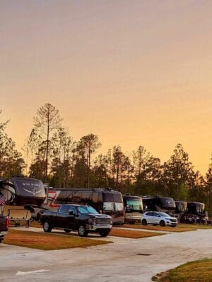 A serene RV campground at sunset, named Savannah Lakes RV Resort, with various RVs and parked vehicles along a paved road. Tall trees frame the background, their silhouettes contrasting against the colorful, fading sky. The warm light creates a peaceful, inviting atmosphere for its grand opening by CRR Hospitality.