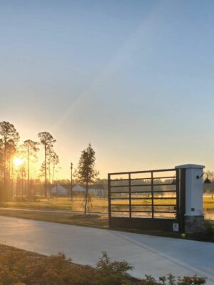 A gated entrance to the Savannah Lakes RV Resort is seen in the early morning light. The gate is flanked by two white pillars with the letters "S" and "L" on them. The sun is rising in the background, casting a warm glow over the trees and landscape, heralding its grand opening with CRR Hospitality.