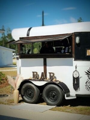A stylish black-and-white mobile bar trailer with a "BAR" sign and a pineapple logo is parked outdoors at the Grand Opening of Savannah Lakes RV Resort. It has rustic charm with minimal decor, including a dreamcatcher and pampas grass. The background features trees and modern buildings under a clear sky.