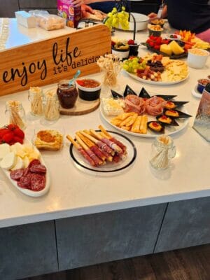 A table at the Savannah Lakes RV Resort Grand Opening is spread with a variety of appetizers, including cheeses, fruits, breadsticks wrapped in ham, and salami. A wooden sign reads "enjoy life, it's delicious." Bowls of dips and spreads, as well as stacks of plates and napkins from CRR Hospitality, are also visible.