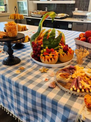 A table with a blue and white checkered tablecloth displaying various foods, including cheese, crackers, fruits arranged in a watermelon basket, bread, and cookies is set up for the grand opening of Savannah Lakes RV Resort. The background shows a modern kitchen with a white countertop and large windows.