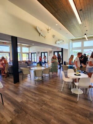 A spacious, well-lit room at Savannah Lakes RV Resort hosts the grand opening event by CRR Hospitality. Some guests are standing and conversing, while others sit at tables adorned with small plants and refreshments. The room features large windows, a wooden ceiling accent, and mounted fish decorations on the wall.
