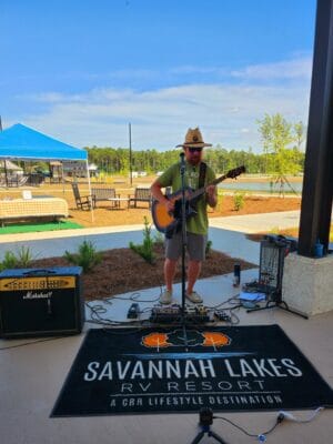 A musician wearing a hat and shorts plays an acoustic guitar and sings into a microphone. He is performing outdoors near a resort pool area. A sign on the ground reads "Savannah Lakes RV Resort: A G&R Lifestyle Destination." Sound equipment is set up around him for the Grand Opening by CRR Hospitality.
