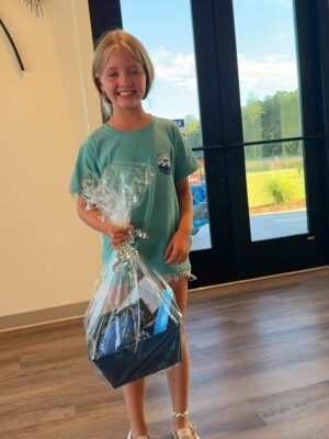 A young girl smiles while holding a blue gift basket wrapped in cellophane. She is wearing a light green t-shirt, denim shorts, and white slip-on shoes. She stands in a brightly lit room at Savannah Lakes RV Resort's Grand Opening, with double glass doors behind her revealing a grassy outdoor area.