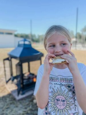 A child with blonde hair enjoys a s'more, standing outside in front of a metal firepit. The child is wearing a white t-shirt with a sun and moon design and the words "COSMIC JOURNEY" printed on it. A clear, blue sky and blurred background of trees and buildings at the Savannah Lakes RV Resort Grand Opening are visible.