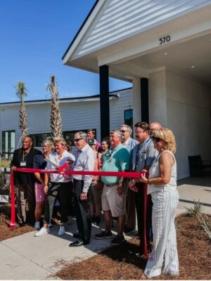 A group of people standing in front of the 570 building, cutting a red ribbon during the grand opening ceremony for Savannah Lakes RV Resort, hosted by CRR Hospitality. The palms and clear blue sky indicate a warm, sunny day.