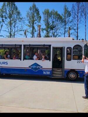 A group of tourists sits inside a white and blue Kelly Tours bus with open windows at the Savannah Lakes RV Resort Grand Opening. A man stands in front of the bus, taking a photo of the passengers. The scene is set outdoors with trees and bushes in the background on a clear, sunny day.