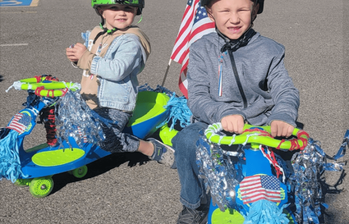 Two children engaged in fun activities wear helmets with colorful decorations as they ride on blue and green toy vehicles. The child on the left sports a spiked helmet and a denim jacket, while the one on the right boasts an American flag and a gray hoodie. Both vehicles are adorned with tinsel and pom-poms.