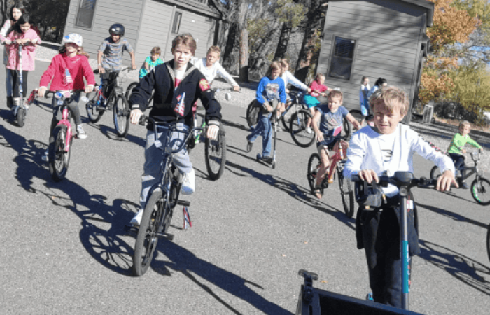 A group of children are enjoying recreational activities, riding bicycles together on a paved outdoor area on a sunny day. Some are ahead while others follow closely behind. Trees and small buildings are visible in the background, adding to the scenic setting.