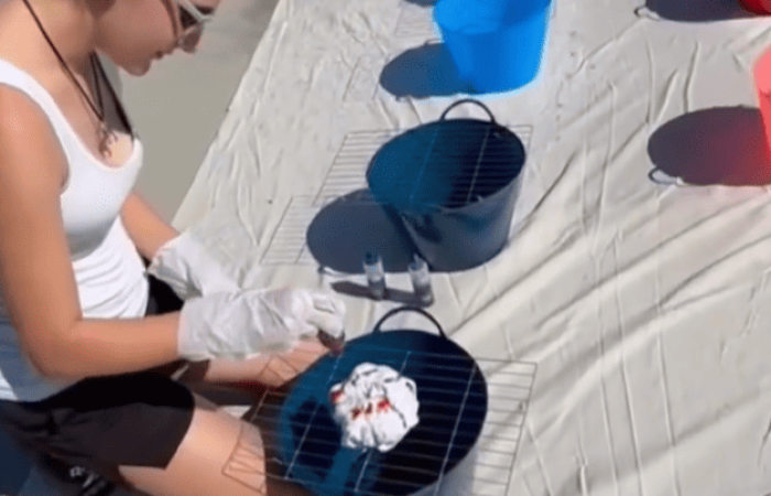 A person with long hair tied back, wearing a white tank top, black shorts, and gloves, is sitting on a large white sheet. They are engrossed in tie-dye activities with white T-shirts and colorful dye bottles, surrounded by several buckets in blue, red, and pink.