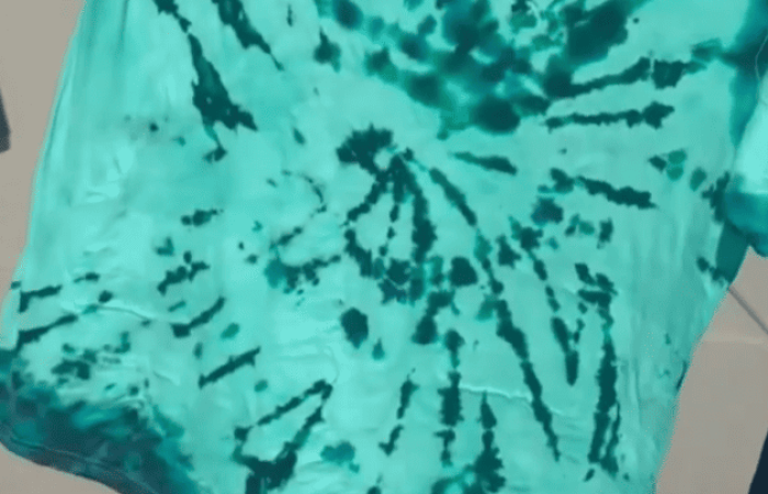 A green and blue tie-dye shirt hanging outdoors. The shirt features radial patterns with various shades of green and blue, creating a vibrant, spiral design. Perfect for outdoor activities, the background shows a sunlit, concrete surface, indicating the image was taken on a bright day.