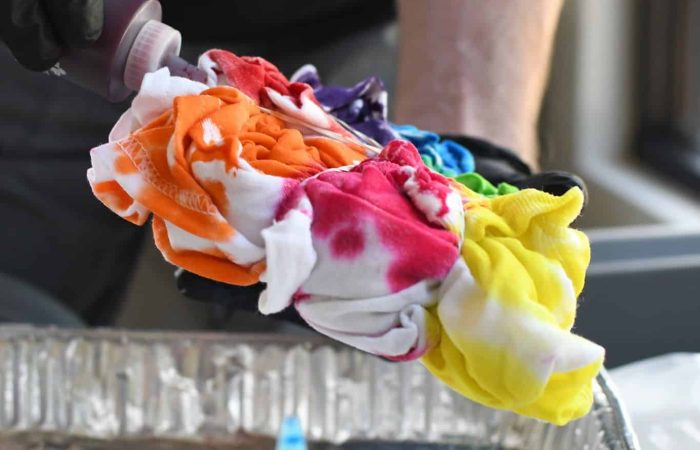 A person wearing black gloves is holding a scrunched-up white fabric being dyed with various colors, including red, orange, yellow, green, blue, and purple. They are applying dye from a bottle over a metal tray during one of those fun activities perfect for creative events like making tie-dye.